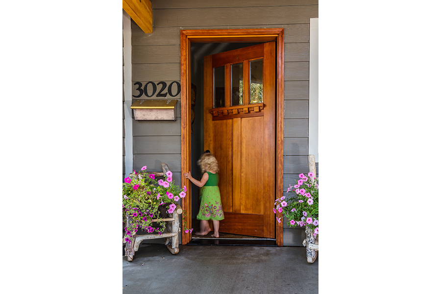 A young curly-haired girl with a green, flower-print dress matching the actual flowers on the porch goes through the front door.